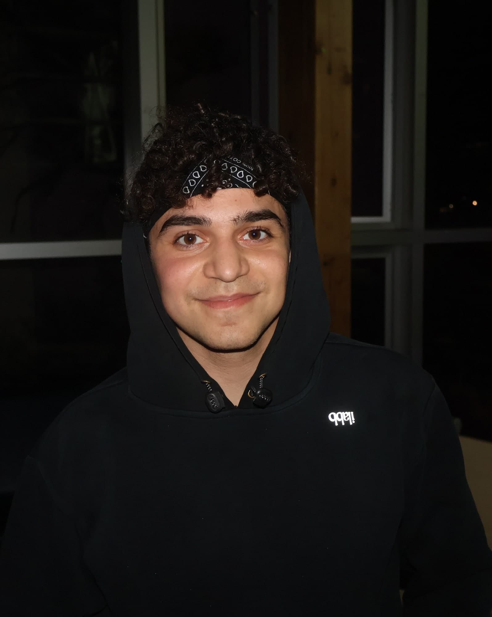 Me smiling in a black hoodie, hood up over a bandana, in front of a window at night